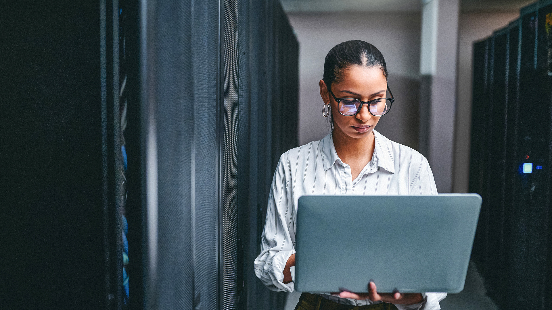 Image of a young IT professional standing in a server room holding a large laptop while researching cyber security in the modern age