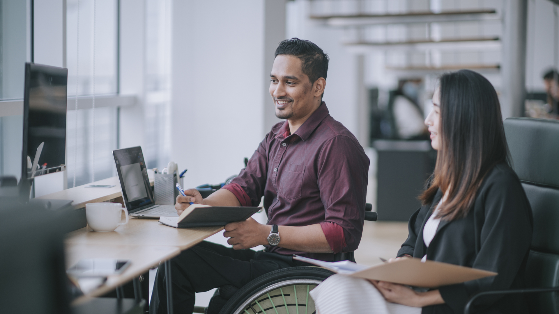 A person in a wheelchair sat next to another person at a desk viewing a digital screen illustrating how managed IT services are helping not-for-profit organisations benefit from tech adoption