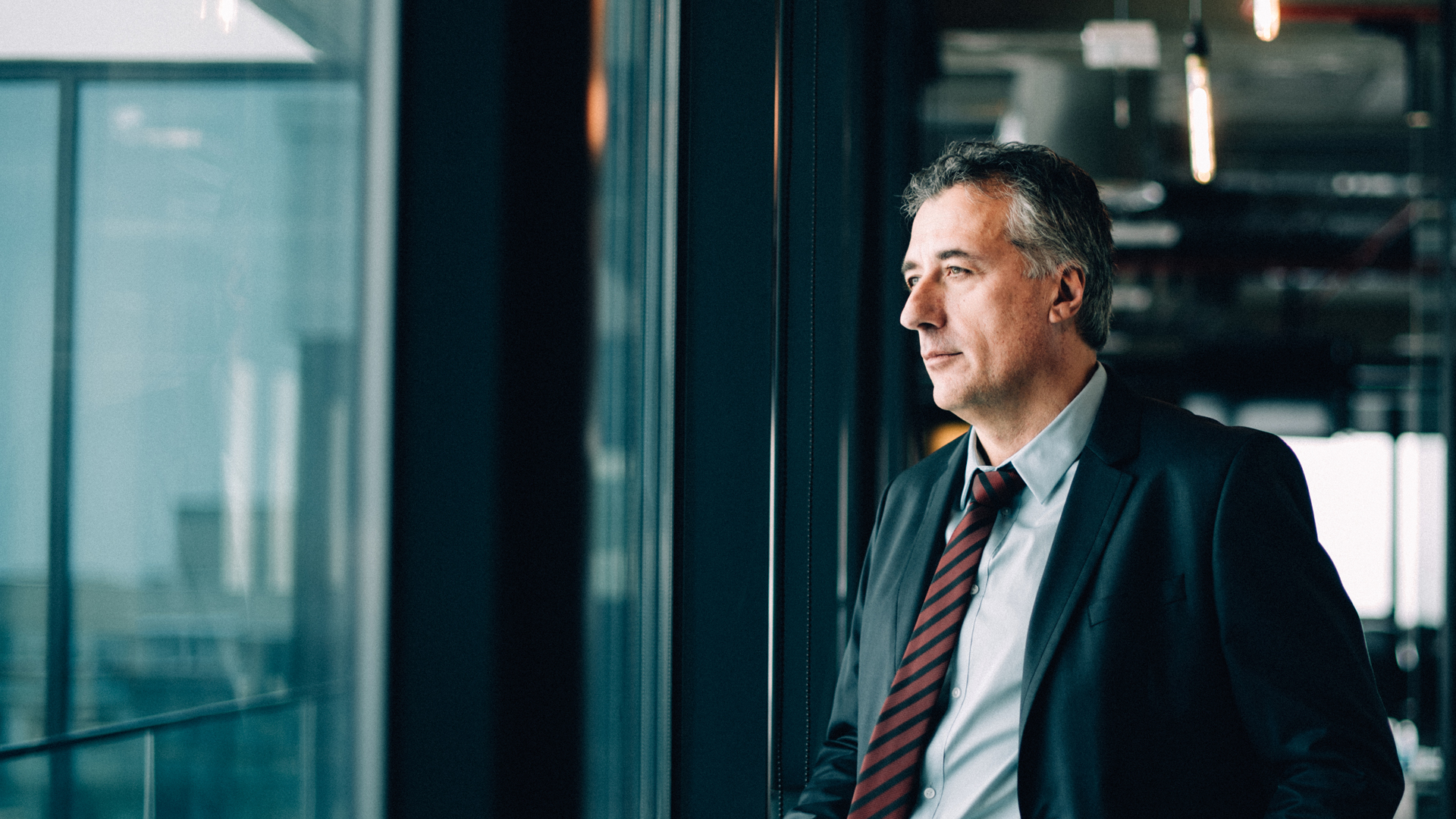 Middle-aged businessman staring out of a glass window in a modern office space while contemplating how to conduct IT audits, and why they are important